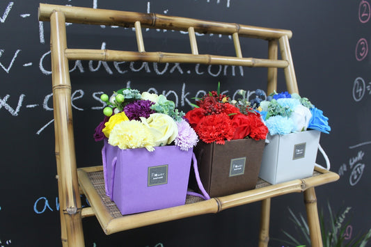 Three small boxes hold colorful artificial flowers on a bamboo shelf against a chalkboard wall reading v-cadeaux.com with circled 2 3 4; box labels read Flower hand made.