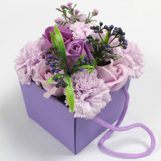 Lavender gift bag contains a compact bouquet of purple roses, carnations, and small berry sprigs, arranged with green leaves, resting on a white surface with a coiled lavender rope handle.