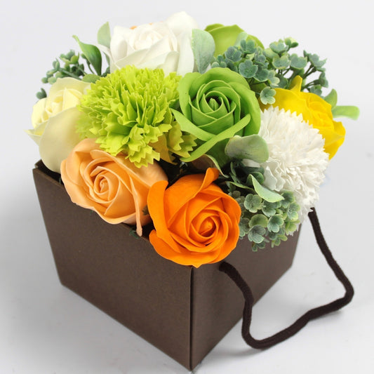 Bouquet of multicolored artificial roses and greenery arranged in a small brown gift bag with dark handles resting on a light surface