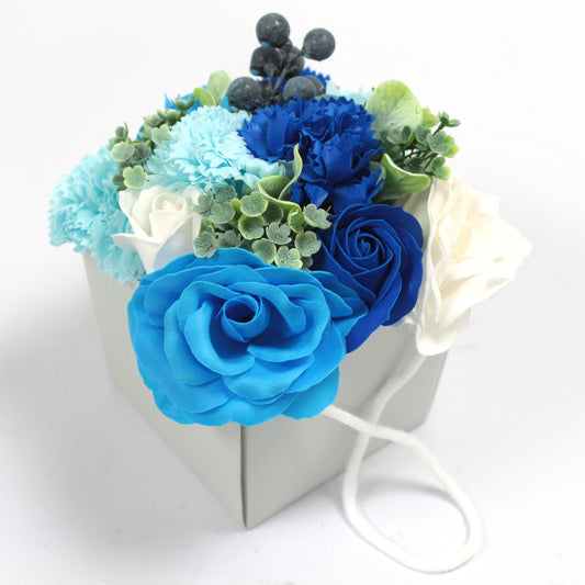 Bouquet of bright blue, navy and white artificial roses and carnations arranged in a small pale gray gift bag with white rope handles on a white surface