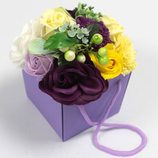 A bouquet of mixed artificial flowers rests in a small purple gift bag, its lavender cord draped on a plain white surface.