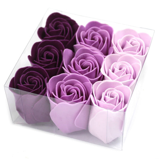 A clear plastic box holds nine artificial roses arranged in three rows by gradient from dark purple to pale lavender on a white background photographed at an angle.