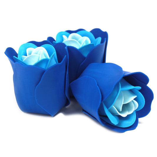 Three blue artificial roses rest closely together on a white surface, two upright and one lying on its side, showing layered petals in deep and light blue tones.