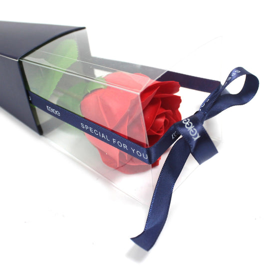 Red rose lies inside a clear plastic gift box tied with a navy ribbon reading "SPECIAL FOR YOU" and "KECE", resting in a navy sleeve on a white background.
