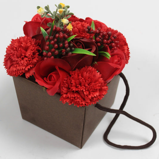 Brown paper gift bag holds a dense arrangement of red roses, carnations and berry sprigs with green leaves, sitting on a white surface with a looped brown rope handle visible.