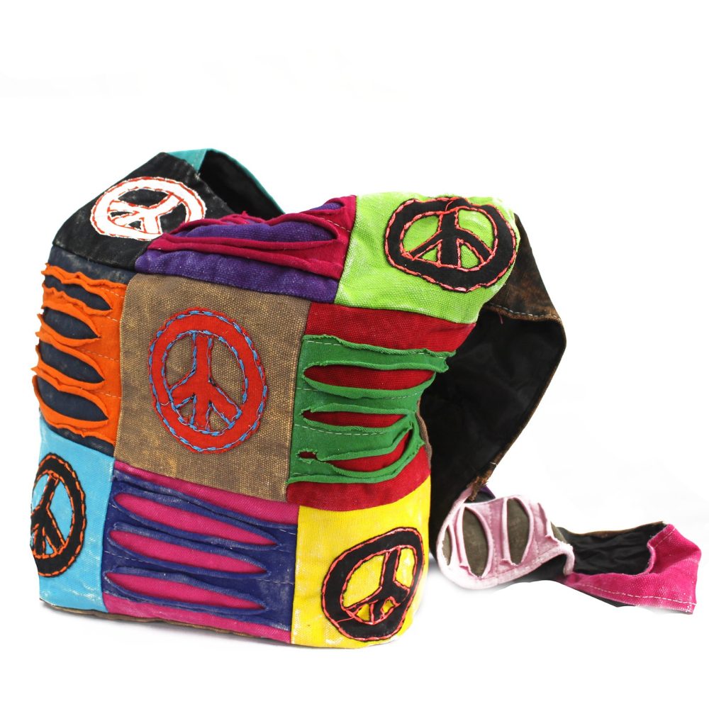 A patchwork-style bag stands upright, displaying various fabric colors and stitched peace symbols, against a white background. Rips and tears expose underlying fabrics, giving a well-worn, bohemian vibe.