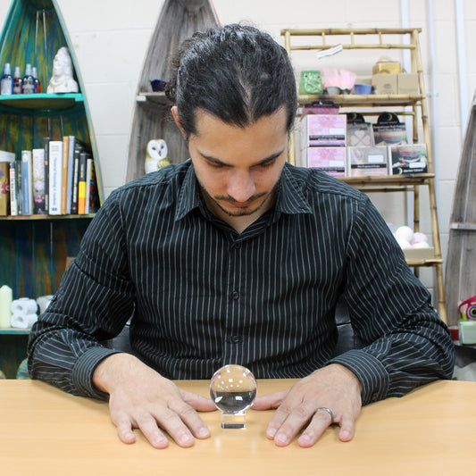 Man concentrates on a crystal ball placed on a wooden table, surrounded by shelves filled with books, candles, and decorative items.
