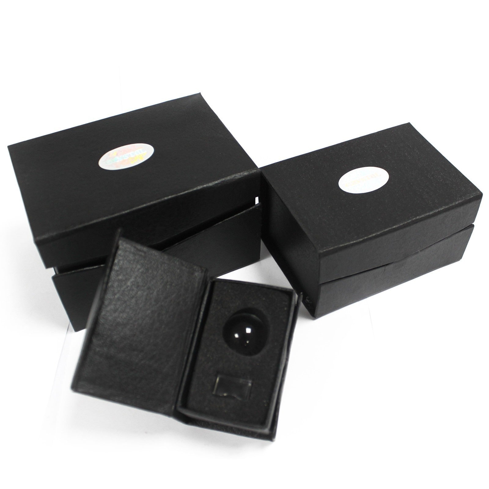 Black boxes with white oval labels are stacked; one box is open, displaying a black spherical object seated in a fitted cushion. The setting is plain and minimalistic.