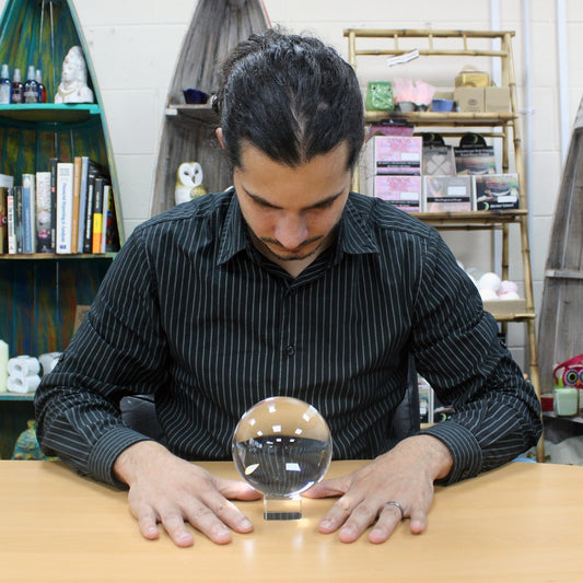 A person with long hair tied back, wearing a striped shirt, gazes at a crystal ball on a wooden table. Shelves with books and decorative items fill the background.