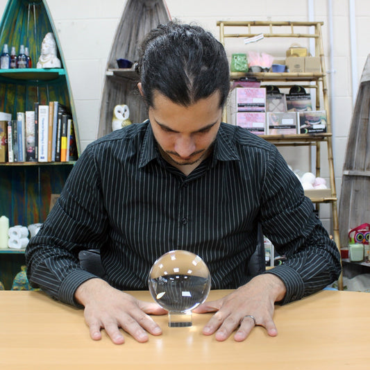 A person with a striped shirt focuses on a crystal ball resting on a table. The background features bookshelves holding various items, including books and decorative objects.