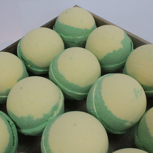 Spherical bath bombs with light yellow tops and green bottoms are arranged closely in a cardboard box, viewed from above in a well-lit setting.