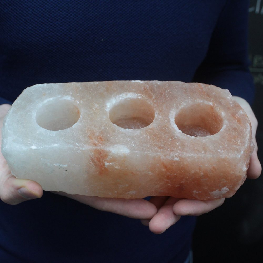 A rectangular, pink-and-white salt rock, carved with three circular recesses, is being held by two hands against a dark background.