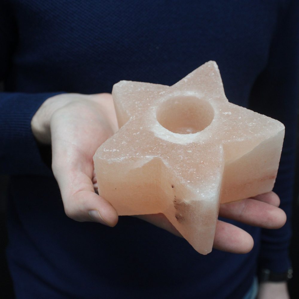 A pink, star-shaped, Himalayan salt candle holder, with a circular indentation on top, is being held in a hand in front of a person wearing a dark blue sweater.