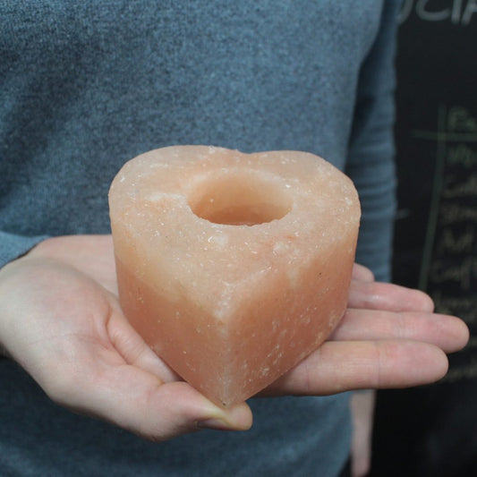 Heart-shaped pink Himalayan salt candle holder rests in open palms of a person wearing a gray sweater, cavity for a candle visible against an indoor dark background with a chalkboard.