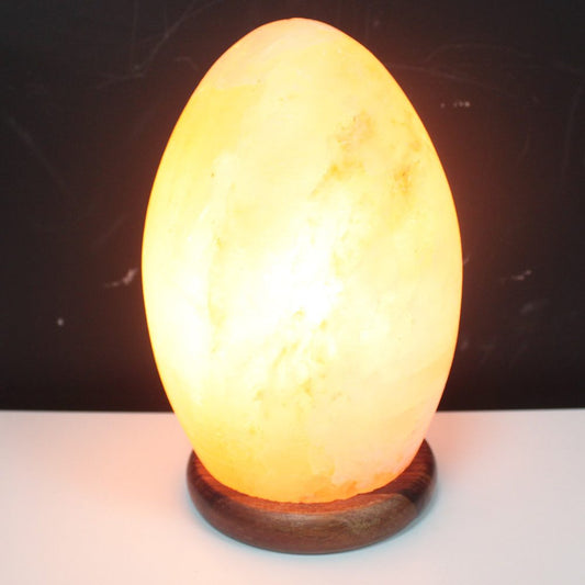 A glowing, egg-shaped salt lamp emits a warm, orange light while resting on a wooden base; set against a dark background on a white surface.