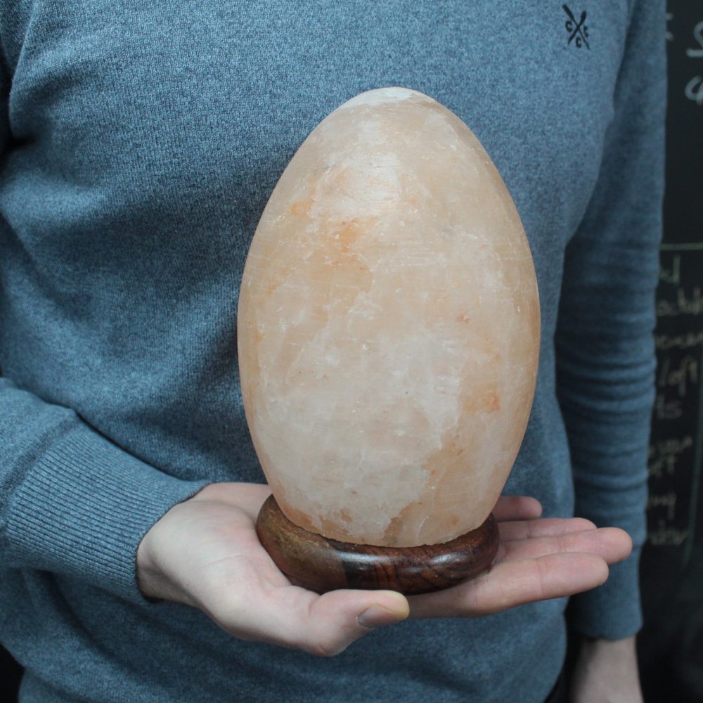 A person holds a large, egg-shaped, orange Himalayan salt lamp on a wooden base, wearing a gray sweater, with a blackboard in the background.