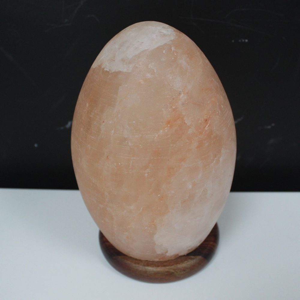 An egg-shaped salt rock sits on a wooden base, resting on a white surface, with a dark background.