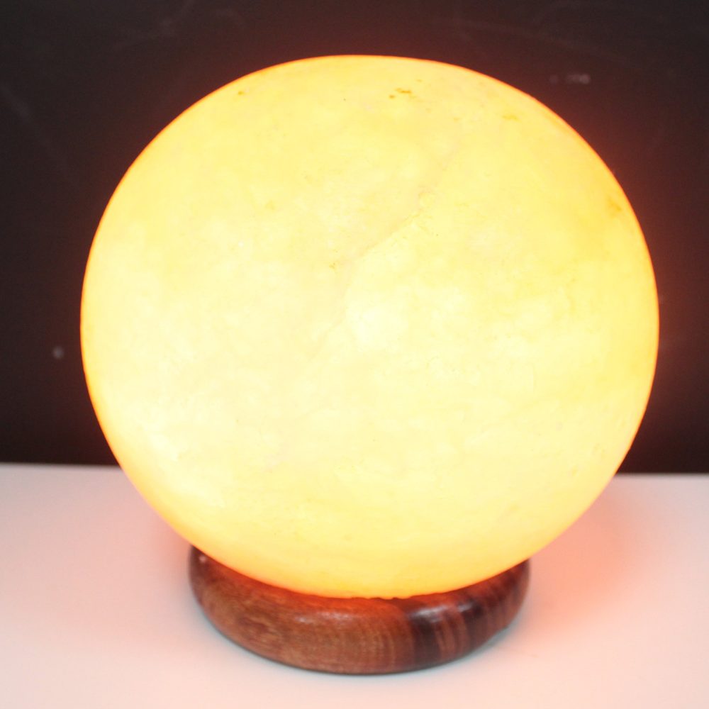 A glowing spherical salt lamp sits on a wooden base, illuminating a soft, warm yellow light in a dimly lit room with a dark background.