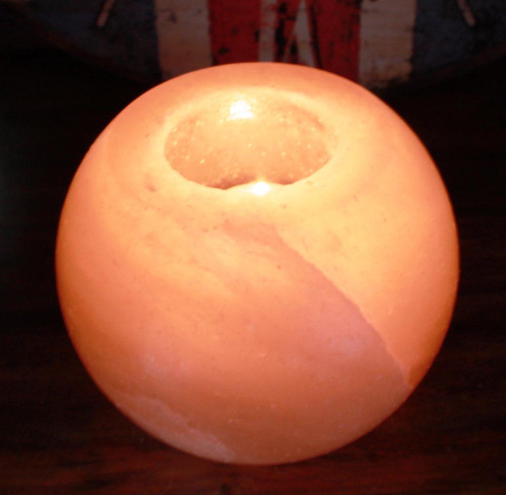 Spherical pink-orange salt candle holder glowing from an inner flame, casting warm light while resting on a dark wooden surface with a shadowed background.