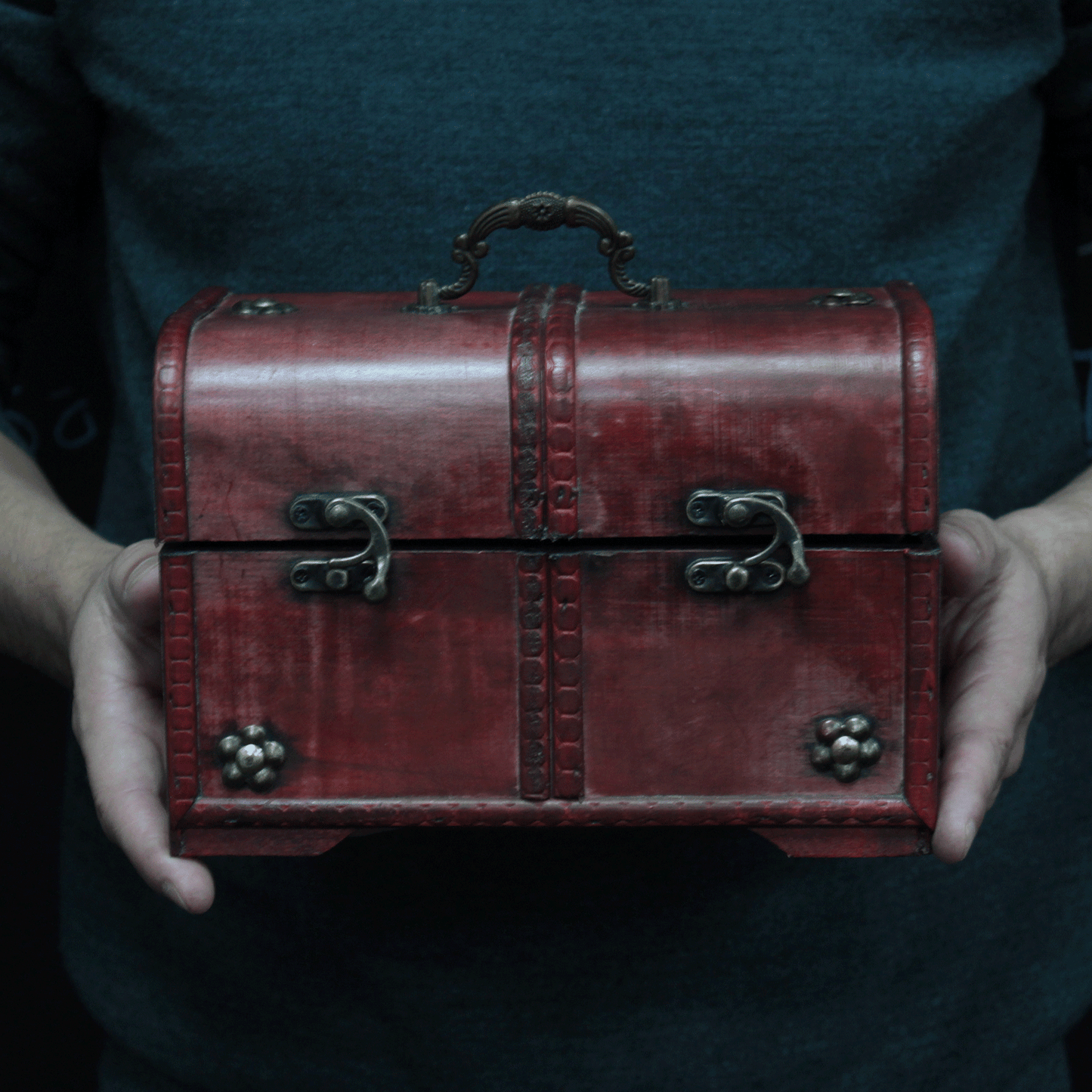 A person holds a small, ornate red chest with metal latches and a decorative handle. The chest has a vintage appearance, against a dark-colored shirt background.