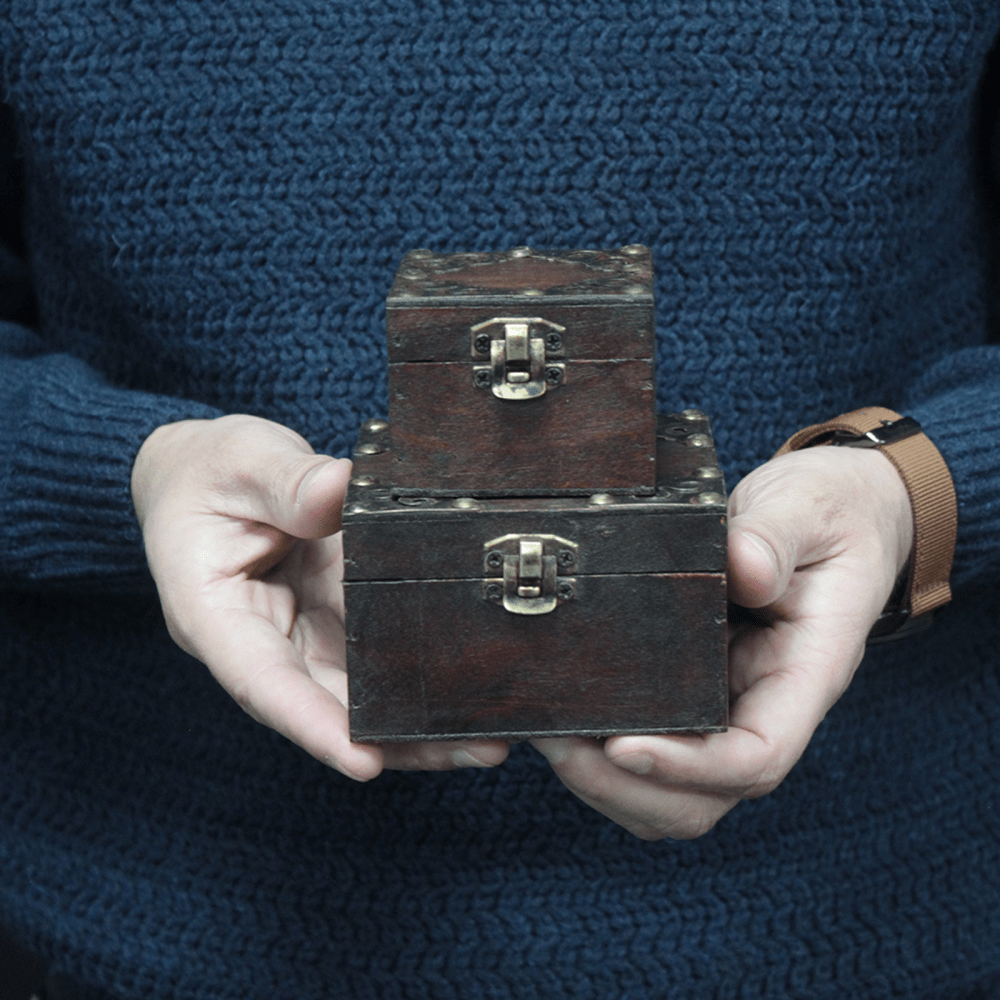 Two small, stacked, wooden chests are held by hands. Each chest features metal latches and decorative studs. The person is wearing a blue knit sweater in the background.