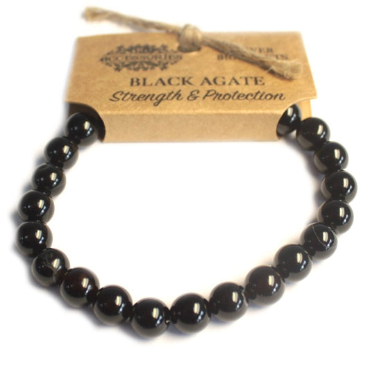A black agate beaded bracelet stretches below a tan tag labeled, "BLACK AGATE Strength & Protection". A twine string hangs off the tag, all resting on a white surface.