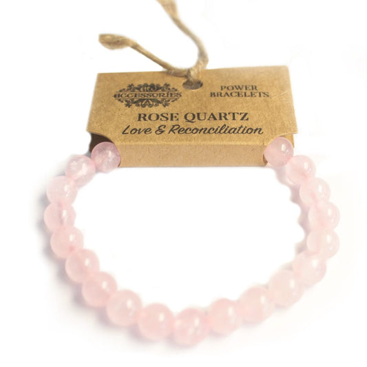A rose quartz bracelet lies in a curve against a white backdrop. The bracelet’s ends are threaded through a tan cardboard tag reading "ACCESSORIES", "POWER BRACELETS," "ROSE QUARTZ Love & Reconciliation" and held by a brown string.