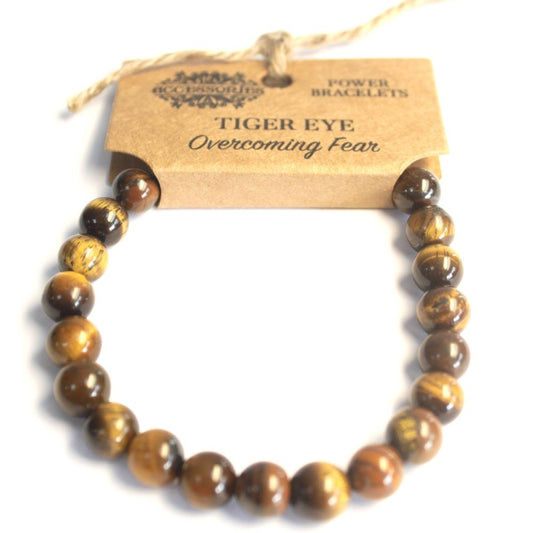 A tiger eye beaded bracelet hangs from a cardboard tag reading "ACCESSORIES POWER BRACELETS TIGER EYE Overcoming Fear," against a white background.