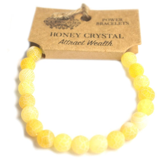 A yellow beaded bracelet with "HONEY CRYSTAL Attract Wealth" printed on a brown tag that reads "ACCESSORIES POWER BRACELETS" sits on a white surface.