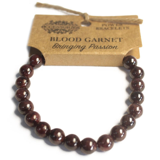 A dark-red bead bracelet forms a circle on a white surface, attached to a tag labeled "ACCESSORIES, POWER BRACELETS, BLOOD GARNET, Bringing Passion" with a brown string.