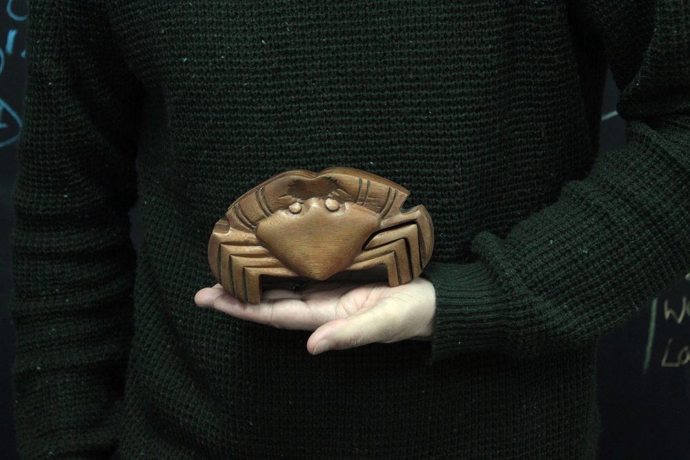 A wooden crab sculpture rests on a person's open palm. The person wears a dark knitted sweater, standing against a dark background with faint chalk markings.