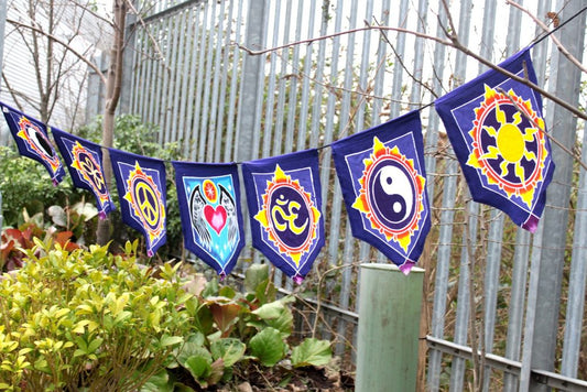 A bunting of dark-blue triangular flags, each displaying a different peace symbol, hangs outside against a metal fence. The symbols include a peace sign, a yin-yang, an Om symbol and a sun.