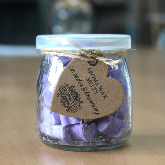 A glass jar filled with purple, heart-shaped wax melts sits on a wooden surface. A heart-shaped tag reads: "AROMA WAX MELTS Lavender & Rosemary" with decorative accents.