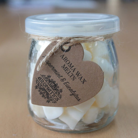 A glass jar contains white wax melts with a heart-shaped brown tag attached by string. Text on the tag reads: "AROMA WAX MELTS Peppermint & Eucalyptus AROMATICS." The jar sits on a wooden surface.