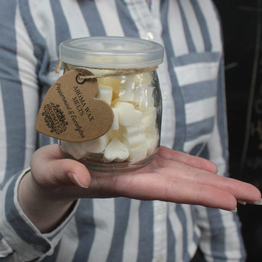 A small glass jar containing heart-shaped wax melts is held in a person's hand. The jar has a label reading: "Aroma Wax Melts Peppermint & Eucalyptus Aromatics".