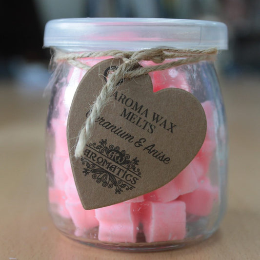 A glass jar contains pink wax melts, labeled with a heart-shaped tag reading "AROMA WAX MELTS Geranium & Anise AROMATICS," tied with a string. It's sitting on a wooden surface.
