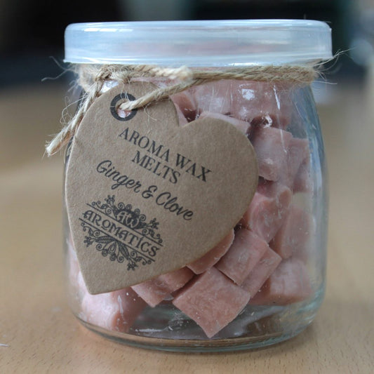 A glass jar contains pink wax melts, with a heart-shaped tag reading, "AROMA WAX MELTS, Ginger & Clove, AROMATICS." The jar is sealed with a tie, set on a wooden surface.