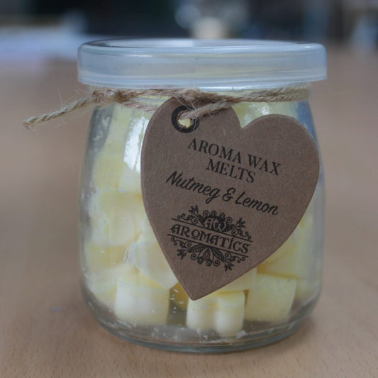 A glass jar filled with yellow aroma wax melts, labeled with a heart-shaped tag: "AROMA WAX MELTS Nutmeg & Lemon AROMATICS," sits on a wooden surface.