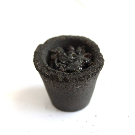 A small, dark, cylindrical container filled with rough, dark granules sits on a white surface, resembling a mini pot with compact contents.