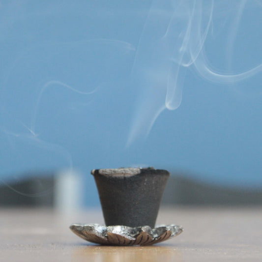 A black incense cone emits curling smoke, resting on a decorative metal holder. The setup is placed on a flat surface, with a blurred blue background.