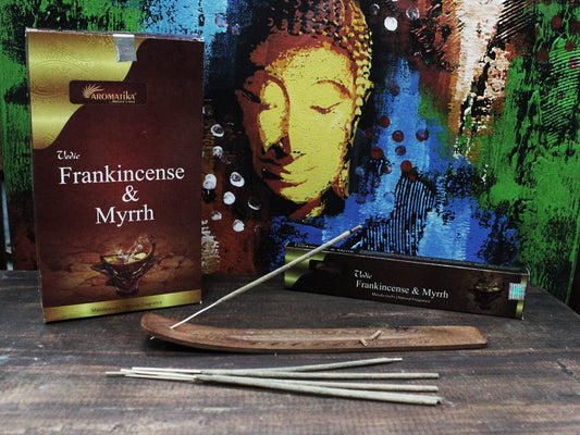 Incense sticks rest on a wooden holder in front of a box labeled "Aromatika, Vedic Frankincense & Myrrh." The background features a colorful abstract painting with a Buddha image.