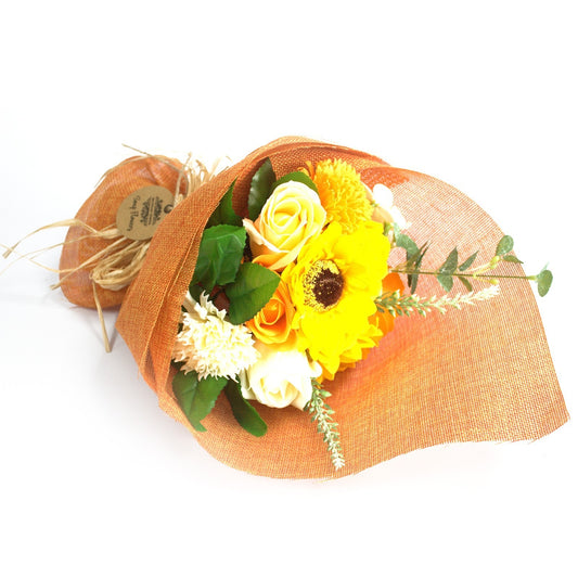 Bouquet of yellow and white flowers wrapped in orange burlap lies angled on a white surface, tied with raffia and a small round kraft tag bearing small unreadable printed text.