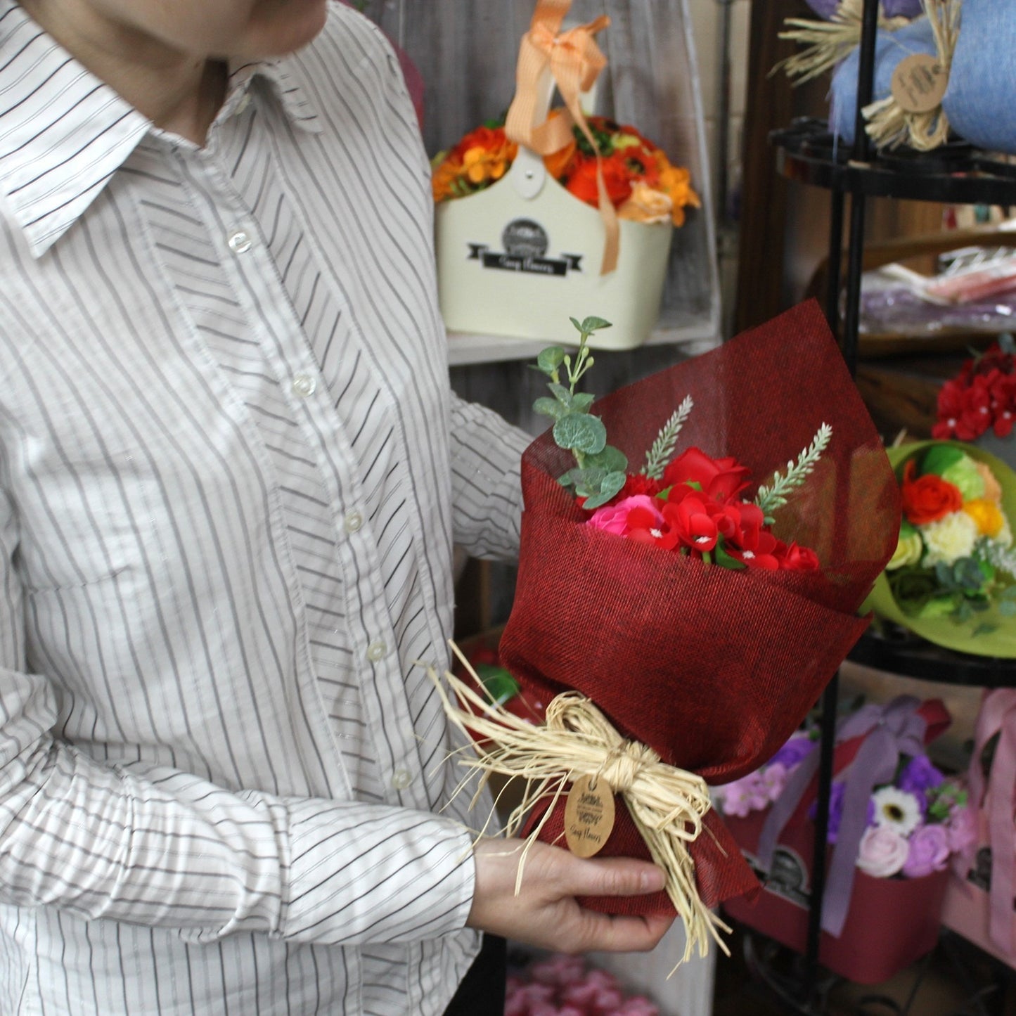 Person wearing striped shirt holds a red-wrapped bouquet of red and pink flowers tied with straw bow while standing in a shop aisle with floral displays; small tag text illegible.