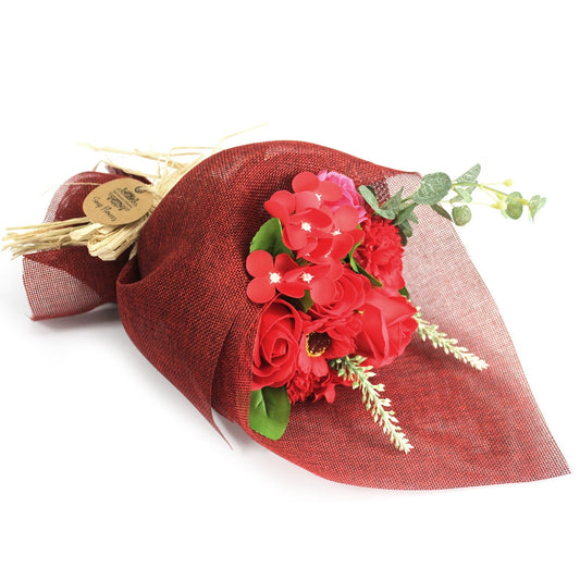 Bouquet of red flowers rests wrapped in burgundy mesh, tied with straw and a small round kraft tag with illegible printed text, placed against a white background.