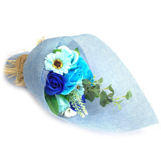 Bouquet of blue and turquoise artificial roses and wildflowers arranged and wrapped in light blue textured fabric, tied with raffia, resting on a white background.