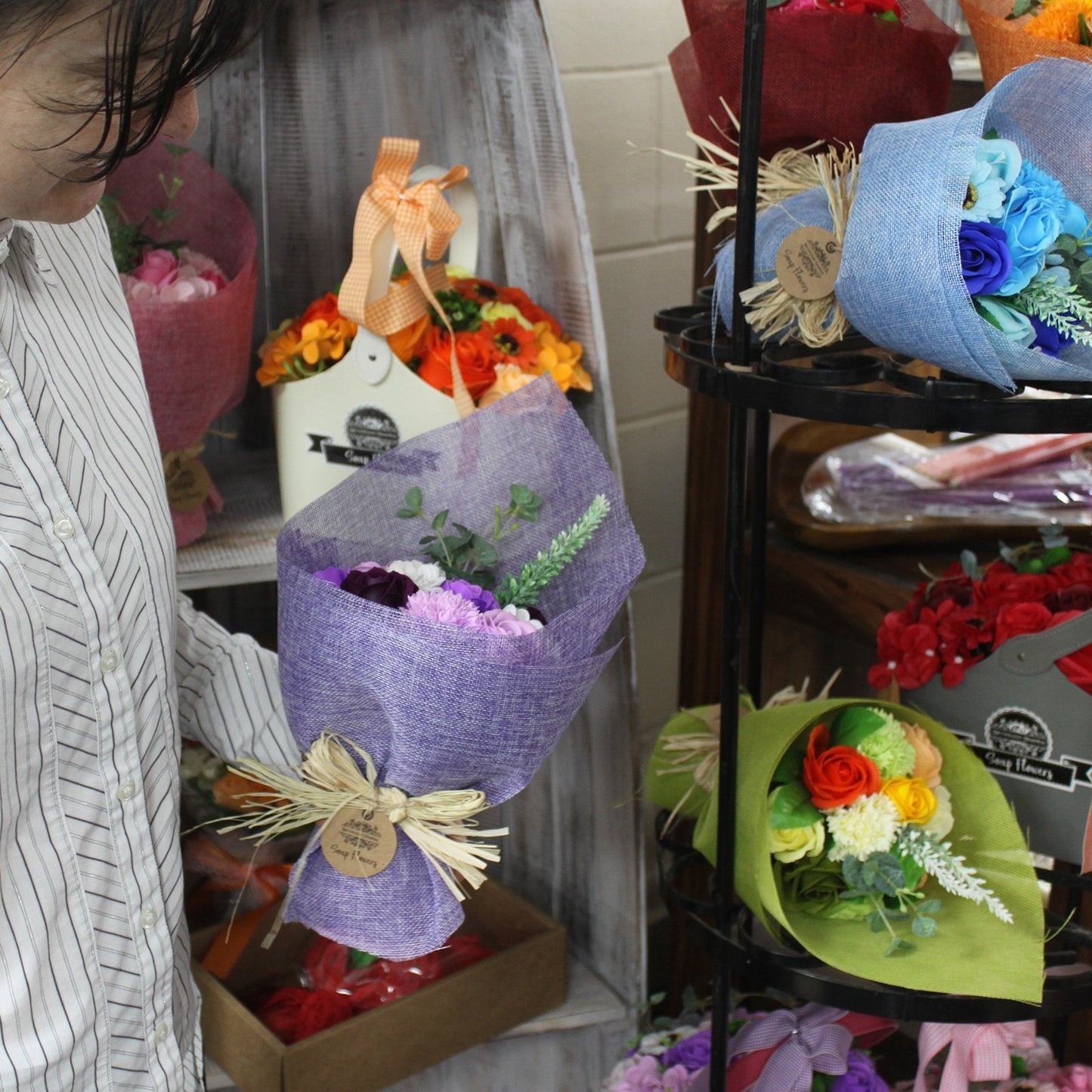 A purple-wrapped floral bouquet is held by a person in a striped shirt amid retail shelving displaying multiple colorful wrapped flower arrangements in a shop.
