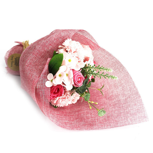 Bouquet of pink artificial roses and mixed blossoms wrapped in textured pink burlap, resting on a white surface with green foliage peeking from the wrap.