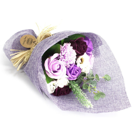 Bouquet of purple, lavender and white artificial roses and greenery rests wrapped in textured lavender fabric tied with a straw ribbon against a white background.