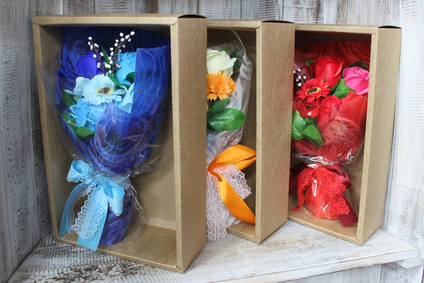 Three small bouquets wrapped in blue, orange, and red, tied with lace ribbons, held upright in cardboard display boxes on a whitewashed wooden shelf.