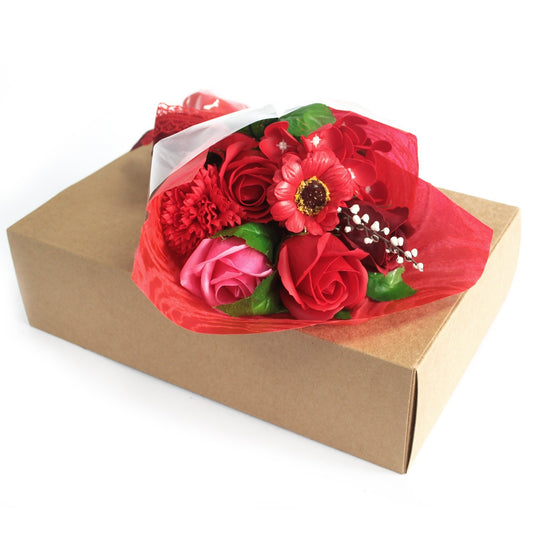 Bouquet of red and pink flowers rests wrapped in red tissue on a closed brown cardboard box against a white background.